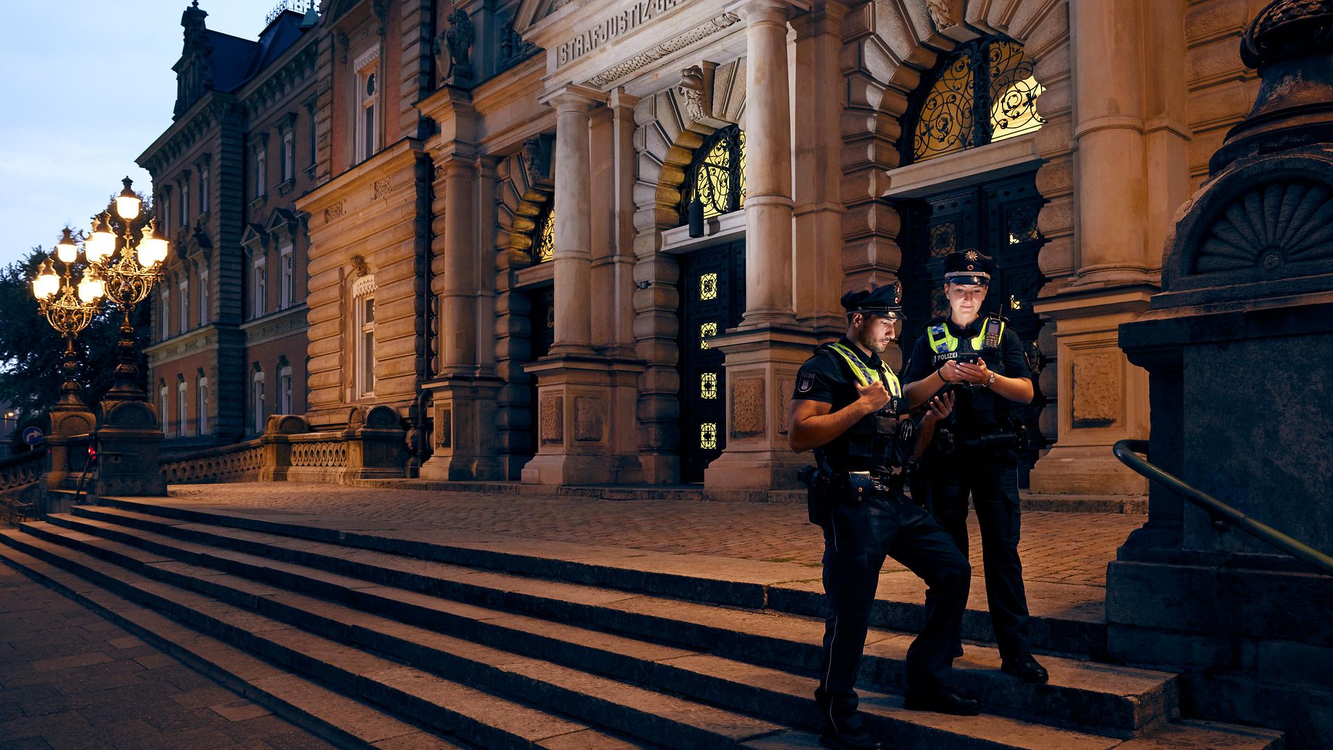 Ein Polizist und eine Polizistin stehen vor dem Strafjustiz-Gebäude und schauen auf digitale Geräte