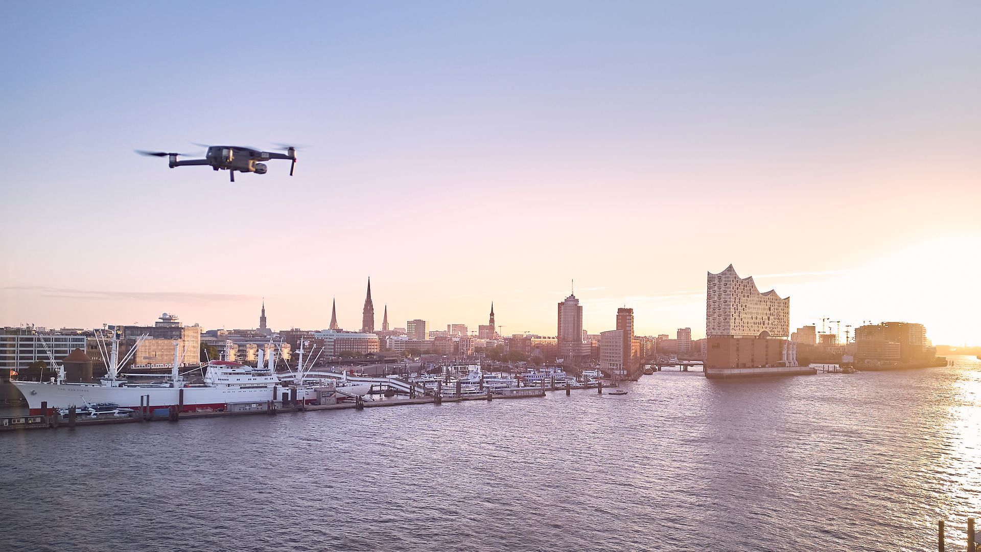 Eine Drohne fliegt über die Elbe im Sonnenaufgang. Elbphilharmonie ist rechts im Bild.